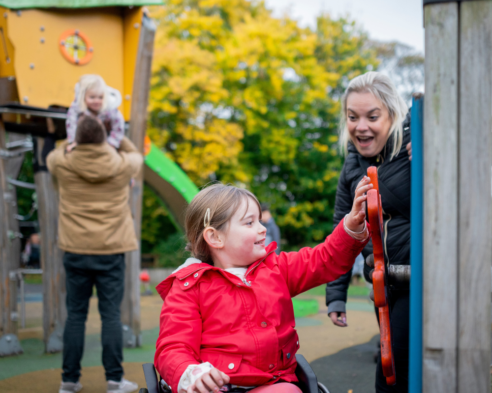 lahdeleikkipuistoon_cropped Perhe leikkipuistossa. Etualalla pyörätuolia käyttävä lapsi pyörittää puista leikkitelineen ruoria vanhemman seuratessa vieressä. Taustalla toinen vanhempi nostaa pienempää sisarusta liukumäkeen.